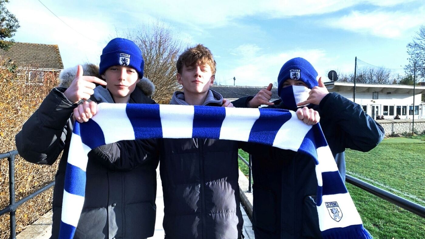 Three young Worcester Park FC supporters in club beanies holding up a blue and white WPFC scarf beside the pitch